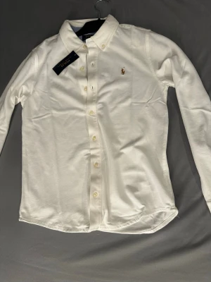 Vit skjorta från Polo Ralph Lauren - Klassisk vit skjorta från Polo Ralph Lauren med broderad björn på bröstet. Skjortan har knappar framtill, lång ärm och krage. Tillverkad i bomull för en bekväm känsla och stilren look.storlek xs passar personer som är ungefär 170