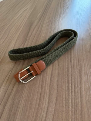 Olivgrönt flätat bälte med läderdetalj - Snyggt olivgrönt bälte i flätad textil med bruna läderdetaljer och silvrigt spänne. Perfekt accessoar för att lyfta din outfit. Längd 115 cm.