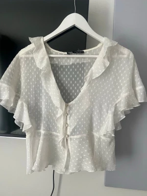 Vit volangblus från Zara - Söt vit blus från Zara med korta volangärmar och djup v-ringning. Blusen är transparent med prickigt mönster, knappar framtill och volangdetaljer längs halsringningen och ärmarna. Perfekt för en trendig och luftig look.