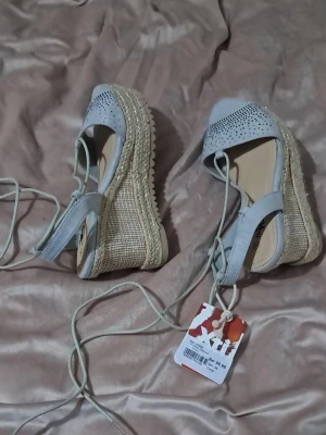 Blå/grå kilklackade espadrilles med snörning - Säljer ett par ljusblågrå espadrilles från XTI med kilklack i flätad jute och öppen tå. Ovandelen har små glittriga stenar och snörning runt vristen för en snygg look. Perfekta till sommaren och ger extra höjd med sin stabila platåsula.