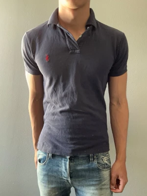 Mörkblå pikétröja från Ralph Lauren - Klassisk mörkblå pikétröja från Ralph Lauren med röd broderad logga på bröstet. Tröjan har krage, korta ärmar och knäppning framtill. Perfekt för en stilren och avslappnad look.