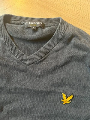 Svart Lyle & Scott tröja med V-ringning - Svart tröja från Lyle & Scott med klassisk V-ringning och gul broderad logga på bröstet. Långärmad modell med ribbade muddar och nederkant. Perfekt för en stilren och enkel look.