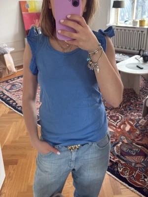 Blå linnetopp med volangärmar - Snygg blå linnetopp med volangdetaljer på ärmarna. Toppen har rund halsringning och är ärmlös med feminina volanger som ger en extra touch. Perfekt till jeans eller kjol för en chill look. Materialet känns mjukt och skönt mot huden.