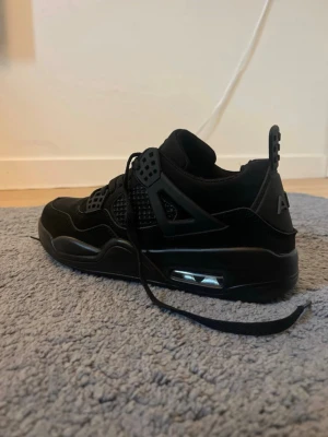 Svarta Air Jordan 4 Retro sneakers - Snygga helsvarta Air Jordan 4 Retro sneakers med meshpaneler, detaljer i plast och synlig Air-enhet i sulan. Skorna har en chunky siluett, klassisk snörning och AIR-logga bak. Perfekta för dig som gillar streetwear och vill sticka ut med en ikonisk modell.