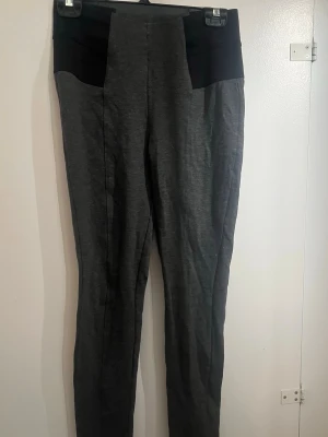 Mörkgrå leggings med svarta detaljer - Snygga mörkgrå leggings med svarta paneler upptill. Hög midja och smal passform som sitter tajt längs benen. Perfekta för en trendig och bekväm stil. Materialet känns mjukt och stretchigt.