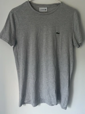 Grå t-shirt från Lacoste - Snygg grå t-shirt från Lacoste med klassisk rund hals och liten broderad krokodil-logga på bröstet. T-shirten är enkel och stilren, perfekt till jeans eller shorts. Tillverkad i mjuk bomull för skön känsla hela dagen.