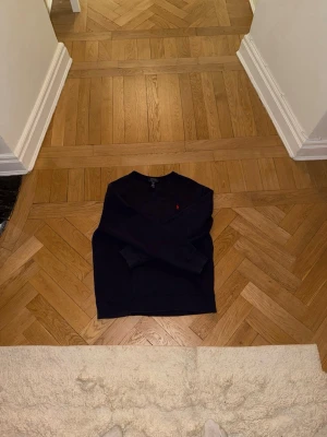 Mörkblå sweatshirt från Polo Ralph Lauren - Mörkblå sweatshirt från Polo Ralph Lauren med klassisk rund hals och diskret röd logga broderad på bröstet. Tröjan är i mjukt bomullsmaterial och har ribbade muddar vid ärmslut och nederkant. Perfekt för en avslappnad och stilren look.
