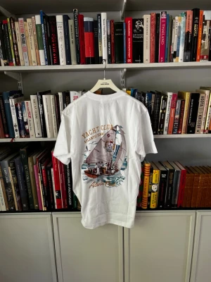 Julian Vane Vit Yacht Club t-shirt  - Helt oanvänd. Vit t-shirt med färgglatt tryck på ryggen som visar en segelbåt och texten 'Yacht Club Atlantic Team'. T-shirten har klassisk passform och korta ärmar, perfekt för en avslappnad och sportig stil.