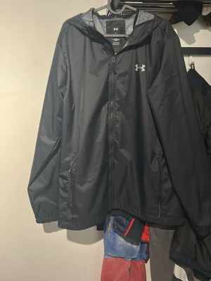 Svart vindjacka från Under Armour - Snygg svart vindjacka från Under Armour med huva och dragkedja framtill. Jackan har två sidofickor med dragkedja och meshfoder på insidan för extra ventilation. Perfekt för blåsiga dagar och sportiga outfits.