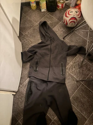 Svart Nike tech - Mysigt svart loungewear set från Nike med matchande hoodie och byxor. Hoodien har dragkedja, huva och två fickor med dragkedja. Byxorna har resår i midjan och sidofickor. Perfekt för chill dagar hemma eller när du vill vara bekväm. Storlek 154-164