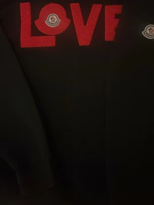 Svart Moncler hoodie med röd LOVE-text - Snygg svart hoodie från Moncler med röd LOVE-text och Moncler-logga på bröstet. Klassisk passform med ribbade muddar och mjukt material. Perfekt för dig som vill sticka ut med en exklusiv streetstyle-look.