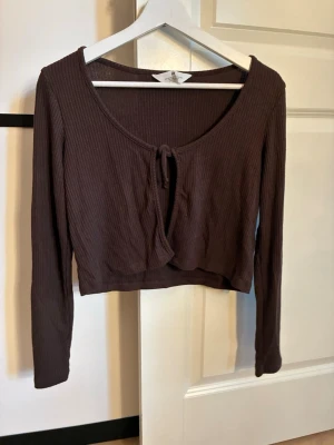 Brun ribbad croppad topp H&M - Snygg brun croppad topp från H&M med långa ärmar och ribbad struktur. Toppen har en söt knytning framtill och är perfekt för att skapa en chill och trendig look. Passar dig som gillar enkla men stilrena plagg.