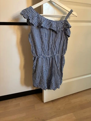 Blårutig jumpsuit med volang H&M - Supersöt blårutig jumpsuit från H&M med volangdetalj och en axelband. Resår i midjan för skön passform. Perfekt för sommardagar och enkel att styla med sneakers eller sandaler. Tunt och luftigt material som känns bekvämt hela dagen.