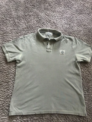 Stone Island piké - Snygg grå pikétröja från Stone Island med klassisk krage och korta ärmar. Tröjan har en broderad logotyp på bröstet och är tillverkad i bomull för en skön och avslappnad känsla. Dessvärre så var den i storlek L men passar perfekt på en som har S! Bara fråga om det är något 