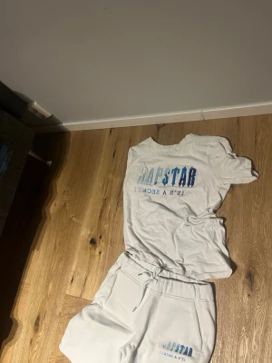 Vit t-shirt och shorts set Trapstar - Säljer ett vitt set från Trapstar med t-shirt och shorts. Båda plaggen har blå och silverfärgad text med loggan 'Trapstar It's A Secret'. T-shirten har rund hals och shortsen har dragsko i midjan. Perfekt för en avslappnad och trendig stil. Storlek xs