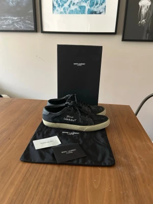 Svarta Saint Laurent sneakers - Svarta sneakers från Saint Laurent med vit broderad logga på sidan. Skorna har en klassisk låg modell, svarta skosnören och en vit, något smutsad sula. Tillverkade i mocka och läder för en lyxig känsla. Perfekta för dig som gillar stilrena och exklusiva sneakers.