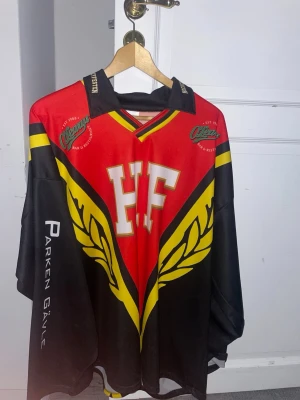 Hockeytröja HF röd/svart/gul - Snygg hockeytröja i rött, svart och gult med stora HF-bokstäver på bröstet och ryggen. Tröjan har tryckta detaljer som O'Learys-logga och Parken Gävle på ärmen. Tillverkad i polyester med klassisk v-ringad krage och långa ärmar. Perfekt för match eller supporterhäng.