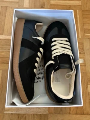 Svarta maison margiela gats storlek 43 - Svarta sneakers från Maison Margiela med vita snören och beige sula. Skorna har en klassisk siluett med paneler i både mocka och läder, samt vit insida. Perfekta för en clean och stilren look.