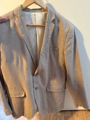 Linne Beige kavaj  - Stilren beige kavaj i mjukt tyg med två knappar framtill, klassisk krage och tre fickor. Fodrad insida och rak passform ger en clean look. Perfekt för dig som vill ha en enkel men snygg kavaj till garderoben.