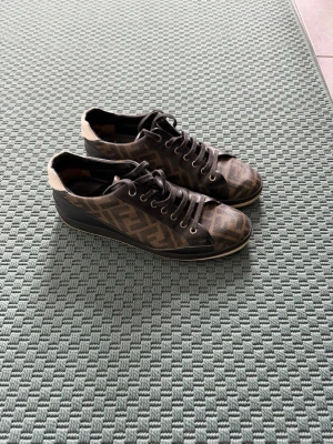 Bruna Fendi sneakers med monogram - Snygga bruna sneakers från Fendi med klassiskt FF-monogram i brunt och beige. Skorna har svarta snören, svarta detaljer och beige hälparti. Tillverkade i canvas och läder för en lyxig känsla och stilren look.