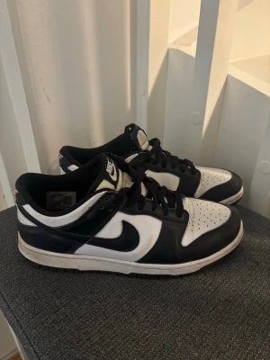 Nike Dunk Low svart/vit sneakers - Snygga Nike Dunk Low sneakers i svart och vitt läder med klassisk swoosh på sidan. Skorna har lågt skaft, svart snörning och perforerad tå för extra ventilation. Perfekta för dig som gillar streetwear och sportig stil.