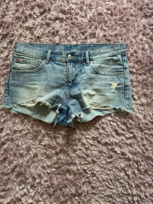 Blå jeansshorts med slitna detaljer - Säljer ett par ljusblå jeansshorts från gina tricot med slitna detaljer och fransig kant. Klassisk femficksmodell med knapp och dragkedja framtill. Perfekta för sommaren och ger en avslappnad vibe. Storlekslappen är bortklippt, men strlk 38/40