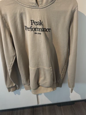 Beige hoodie från Peak Performance - Beige hoodie från Peak Performance med svart broderad logga och text 'Est. 1986' på bröstet. Klassisk känguruficka framtill, ribbade muddar och huva. Perfekt för en avslappnad och stilren look.