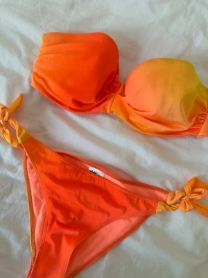 Orange bandeau bikini med avtagbar axelband - Snygg bandeau bikini i orange och gul med knyt på sidorna av underdelen. Toppen är axelbandslös och har en mjuk övergång mellan färgerna. Perfekt för stranden eller poolen. Materialet känns mjukt och stretchigt.  ÖVERDELEN ÄR I STORLEK 80C  OCH UNDERDELEN I STORLEK 36