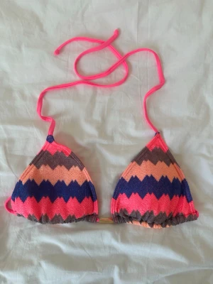 Färgglad zigzag bikiniöverdel - Snygg bikiniöverdel med zigzag-mönster i rosa, blått, orange och brunt. Triangelformade kupor och tunna knytband i neonrosa. Perfekt för stranden eller poolen och ger en färgklick till din sommarstil.
