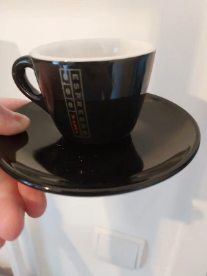 Svart espressokopp med fat - Snygg svart espressokopp med tillhörande fat. Koppen har vit insida och svart utsida med texten 'ESPRESSO' på sidan. Tillverkad i keramik och perfekt för en liten kaffe på morgonen eller efter maten.
