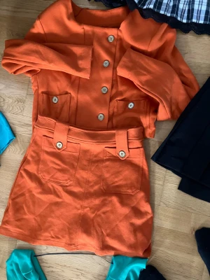 Orange bodyset med knappar - Snyggt orange bodyset med matchande överdel och kjol. Setet har dekorativa träfärgade metall knappar framtill och stora fickor på både överdel och kjol. Stilren design med markerad midja och bältesdetaljer. Perfekt för dig som vill sticka ut med färg.