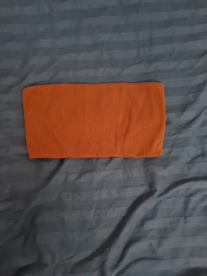 Ribbad orange bandeau topp - Snygg ribbad bandeau topp i orange färg. Toppen är axelbandslös och har en tight passform, perfekt för att styla med höga jeans eller kjol. Materialet känns mjukt och stretchigt för en bekväm känsla.