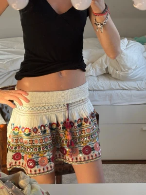 Vit kortkjol med broderade blommor - Supersöt vit kortkjol med färgglada broderade blommor och volangkant. Kjolen har bred smock i midjan och dekorativa tofsar framtill. Perfekt för dig som gillar boho-stil och vill ha något unikt till sommaren. Från en gammal Zara kollektion som inte säljs längre❤️❤️