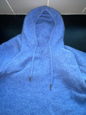 Kashmir Hoodie - En mysig blå hoodie i fluffigt material med huva och dragsko med metalländar. Hoodien har en avslappnad passform och långa ärmar. Perfekt för chill dagar eller när du vill vara extra bekväm.