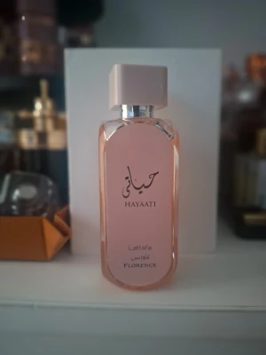 Lattafa Hayaati Florence parfym - Elegant parfymflaska i ljusrosa glas med silverdetaljer och fyrkantigt lock. Flaskan har texten Hayaati Florence på framsidan och utstrålar en modern och stilren känsla. Perfekt för dig som gillar exklusiva dofter och snygg design.