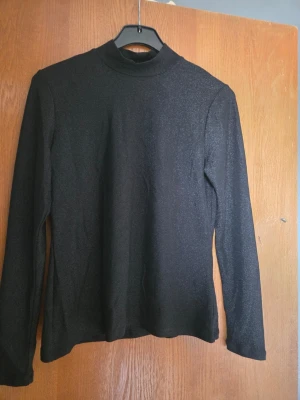 Svart glittrig polotopp - Snygg svart polotopp med glittrigt material och lång ärm. Perfekt för dig som vill ha en enkel men ändå iögonfallande topp. Polokrage och figurnära passform gör den lätt att styla till olika looks.