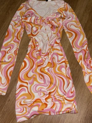 Färgglad swirlmönstrad kortklänning - Säljer en kort klänning med färgstarkt psykedeliskt mönster i rosa, orange, gult och vitt. Klänningen har lång ärm, v-ringning och rynkade detaljer framtill. Perfekt för dig som gillar retrostil och vill sticka ut.