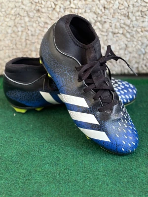 Adidas Predator blå fotbollsskor - Säljer ett par Adidas Predator fotbollsskor i svart och blått med vita detaljer och snörning. Skorna har en högre strumpliknande krage och mönstrad ovansida för bättre bollkontroll. Perfekta för dig som vill sticka ut på planen. Nästan helt nya, de har varit liten och kan diskutera priset vid snabbäfare 