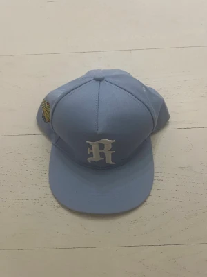 Ljusblå keps med broderad R-logga - Snygg ljusblå keps med vit broderad R-logga framtill och en färgglad patch på sidan. Klassisk form med böjd skärm och justerbar baksida. Tillverkad i bomullstyg som känns bekvämt och lätt på huvudet. Perfekt för att lyfta din streetwear-look.