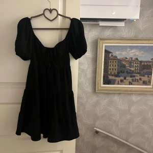 Svart klänning med puffärm - Supersöt svart kortklänning med puffärmar och knyt framtill. Klänningen har en luftig passform och volang i kjolen. Köpt från ginstricot och endast använd 1 gång✨