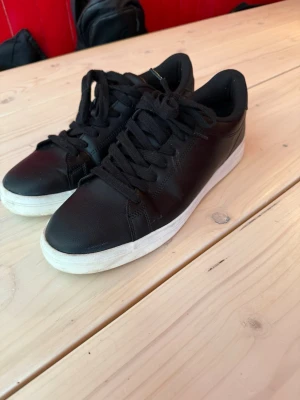 Svarta sneakers med vit sula - Stilrena svarta sneakers med lågt skaft och vit sula. Skorna har svarta skosnören och är tillverkade i slätt syntetmaterial som ger en clean look. Perfekta för dig som gillar enkel och klassisk stil. Storlek 45