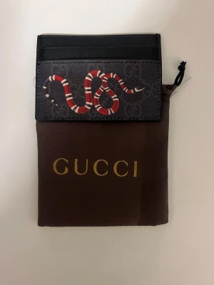 Gucci korthållare med orm-motiv - Snygg korthållare från Gucci i svart och mörkgrått med det klassiska GG-mönstret och en rödvit orm på framsidan. Tillverkad i canvas och läder, perfekt för kort och sedlar. Kommer med originalpåse med guldfärgad logga.