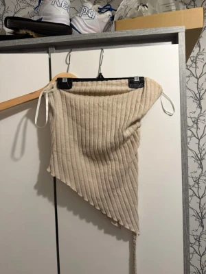 Beige ribbad asymmetrisk tubtopp - Trendig beige ribbad tubtopp med asymmetrisk skärning och snörning i sidan. Toppen är ärmlös och har en croppad passform, perfekt för att styla med höga jeans eller kjol. Materialet känns mjukt och stretchigt, vilket gör den bekväm att bära.