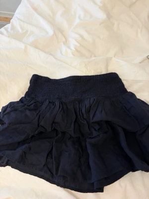 Mörkblå volangkjol från Bershka - Supersöt mörkblå kortkjol från Bershka med bred smock i midjan och flera lager volanger. Kjolen har en flowy och luftig känsla, perfekt för varma dagar. Enkel att matcha med olika toppar för en trendig look.