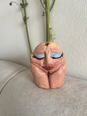 Handgjord skulptur med ansikte - Unik handgjord skulptur i keramik med ett abstrakt ansikte. Skulpturen är i hudfärgad ton med blå ögonlock, svarta detaljer och röda läppar. Formen är rundad och kompakt, med tydliga ansiktsdrag och konstnärlig stil. Perfekt som dekoration eller samlarobjekt.