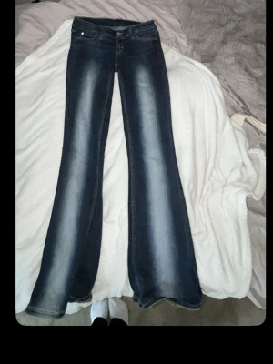 Mörkblå bootcut jeans med slitningar - Säljer ett par mörkblå bootcut jeans med snygga slitningar och ljusare tvätt längs benen. Jeansen har klassiska fem fickor och kontrastsömmar. Perfekta för dig som gillar en retro vibe och vill ha ett par jeans som sticker ut lite extra.