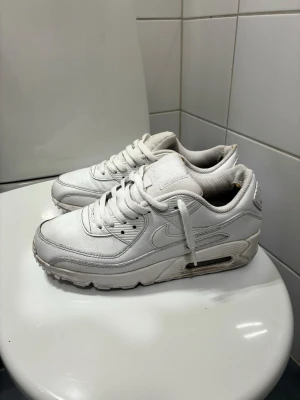 Vita Nike Air Max 90 sneakers - Klassiska vita Nike Air Max 90 sneakers med synlig Air-enhet i sulan. Skorna har en chunky siluett, mesh och läderdetaljer samt vita skosnören. Perfekta för dig som gillar streetwear och sportig stil.