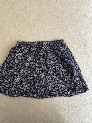 Mörkblå blommig minikjol med knappar - Supersöt mörkblå kortkjol med små vita blommor och knappar framtill. Den är från Stradivarius och är i storlek S.