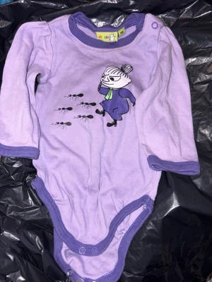 Säljes Lila Mumin body med motiv, strl 56 - Supersöt lila body från Moomin med långa ärmar och lila kantband. Framsidan har ett tryck med Lilla My och myror. Tryckknappar i grenen och för enkel påklädning. Tillverkad i mjukt material, perfekt för de minsta. Katt finns i hemmet. Kan tas med när jag åker mot Nykvarnshållet eller Järna eller mot Stockholmshållet eller också skickas mot fraktkostnad. Säljes för 211kr plus frakt.