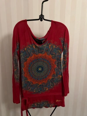 Röd långärmad topp Desigual mönster - Unik röd långärmad topp från Desigual med stort färgstarkt mandalamönster i blått, orange och grönt framtill. Toppen har rund halsringning och dekorativ knytning i nederkant. Perfekt för dig som gillar färg och detaljerad design.